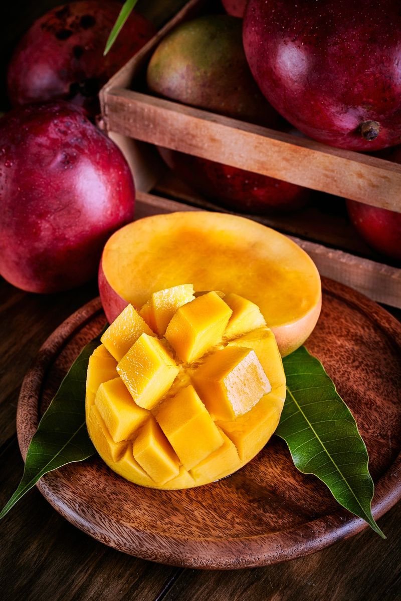 Tropical fruits. Close-up of mango slice with mangos in a crate on rustic table. The mango belongs to the genus Mangifera and to the flowering plant family Anacardiaceae.  The mango is a juicy stone fruit and is native to south and southeast Asia, and now available all over the world. Consisting of numerous tropical fruiting trees and with many varieties it is cultivated mostly for edible fruit. The majority of these species are found in nature as wild mangoes, nevertheless, in South America there are many big plantations for commercial purposes. The mango with the banana has become two of the most cultivated fruits in the tropics