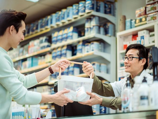 A customer hands a shopping basket to a friendly pharmacist behind a counter.







