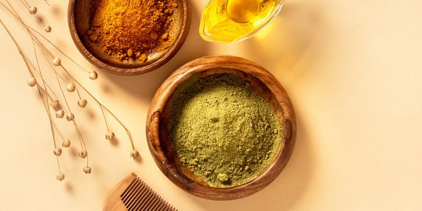 Wooden bowls with orange and green powders, oil, and a wooden comb on a beige background.