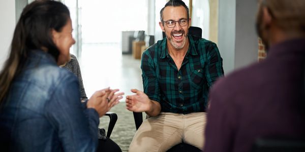 Man in plaid shirt laughing and clapping with friends in a casual meeting.