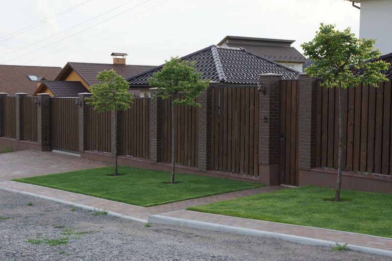 long private brown fence made of wooden boards and bricks with a closed front door on the street with green grass and decorative trees