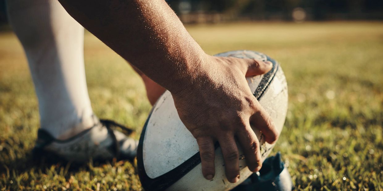 A close-up of a hand holding a rugby ball on a grassy field.
