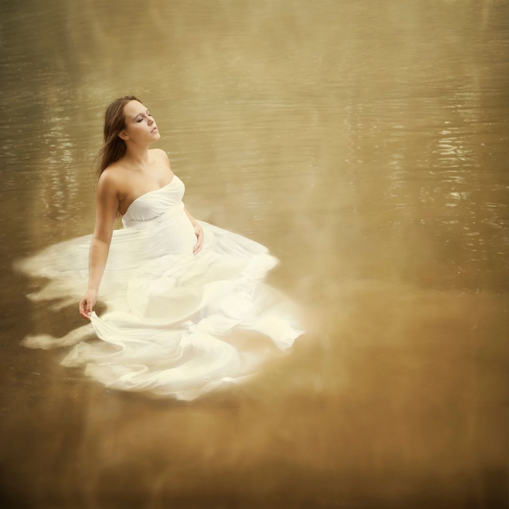 A serene pregnant woman in a flowing white dress sits peacefully in calm water.