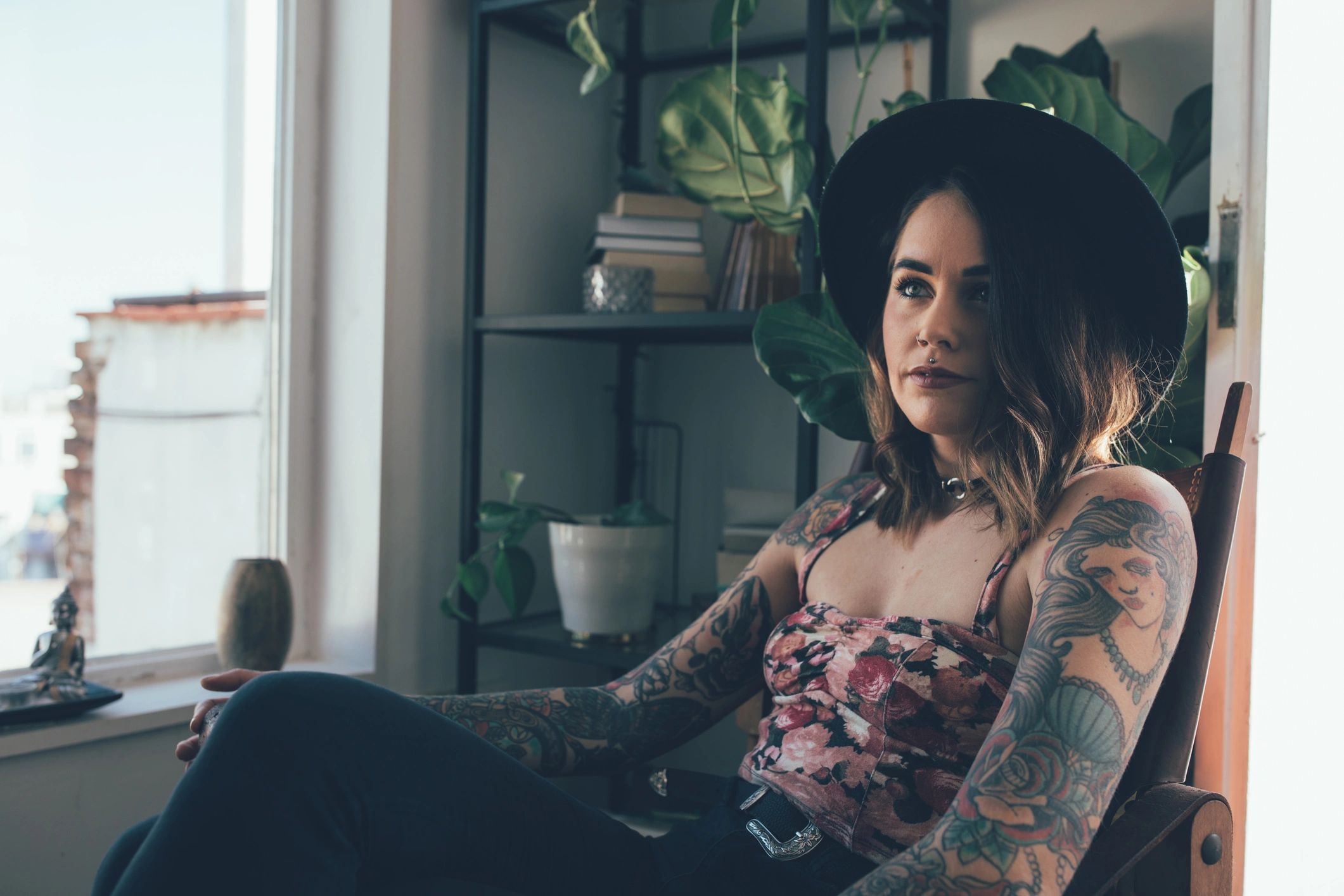 Woman with tattoos and a black hat sitting indoors near a window.