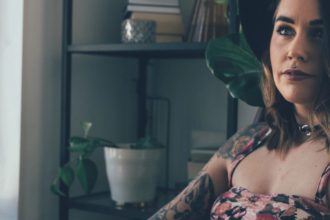 Woman with tattoos and a black hat sitting indoors near a window.