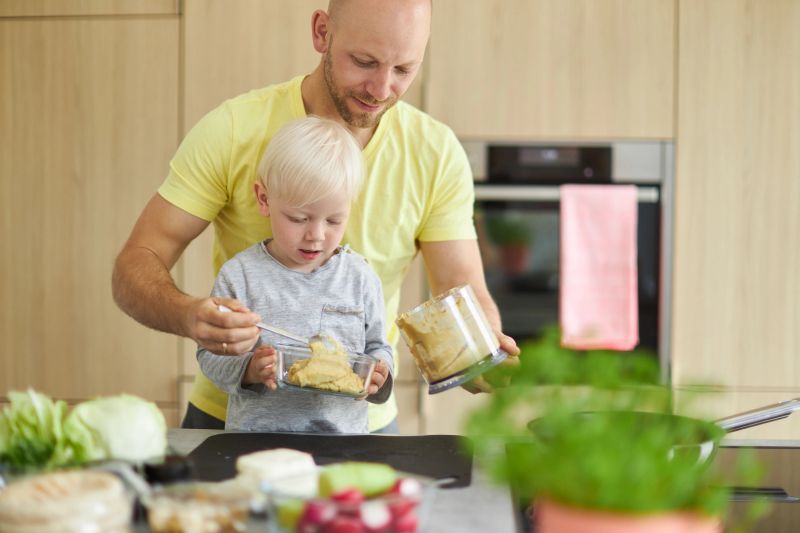A father prepares a sandwich and homemade hummus with his son in the modern kitchen at home while he shows him and explains how to cook, cut vegetables and operate the blender and the garlic press. he wears a yellow t-shirt and have fun