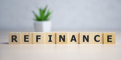 Wooden blocks spell out the word REFINANCE on a table with a plant blurred in the background.