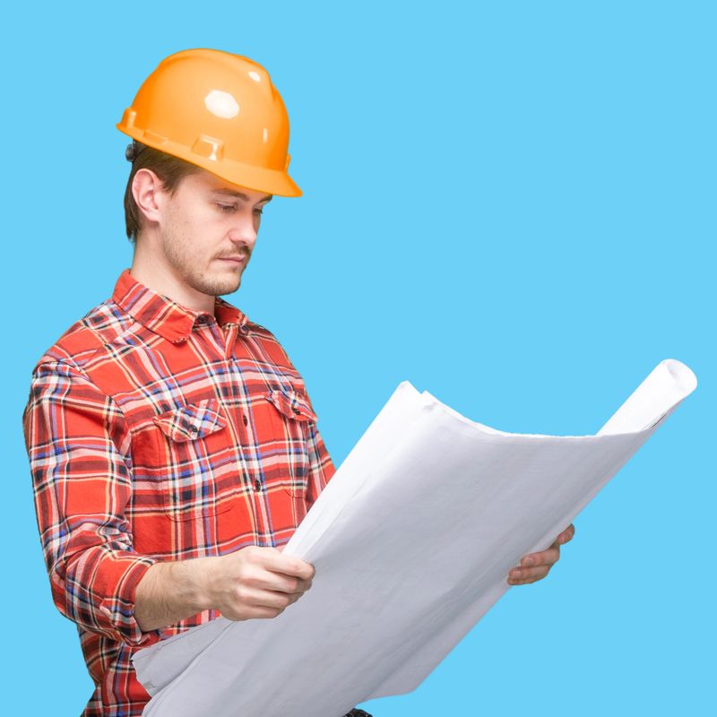 Waist up of with brown hair male construction worker standing wearing hardhat who is learning who is examining and holding hammer and using blueprint