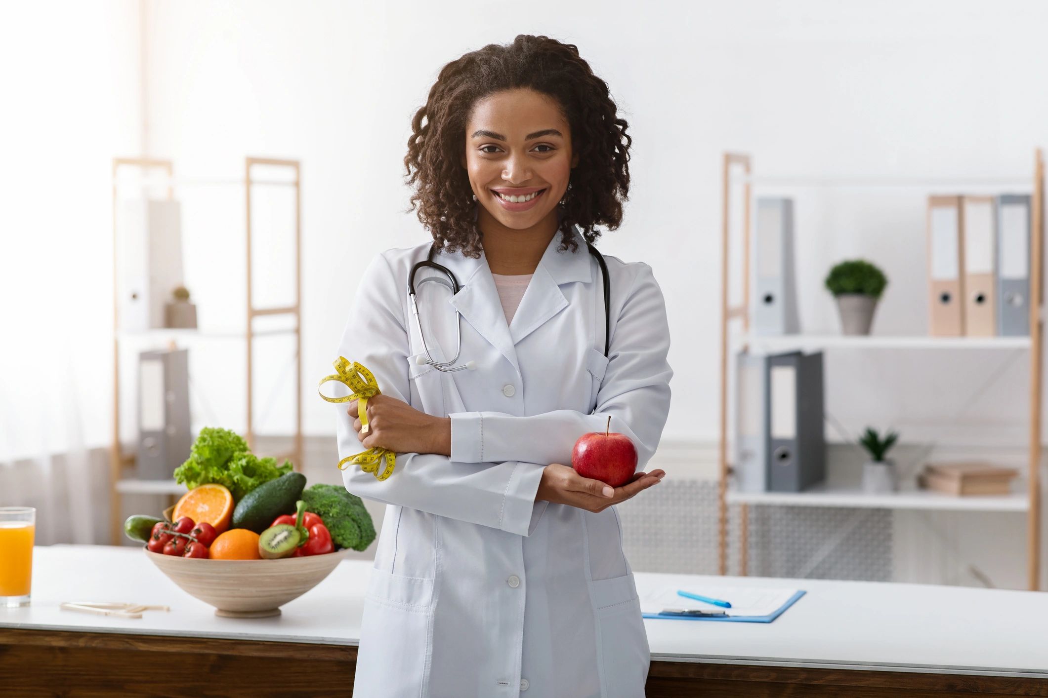 Smiling dietitian holding an apple and measuring tape in a bright office.