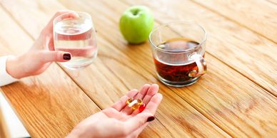 Person holding pills and a glass of water with an apple and tea on the table.