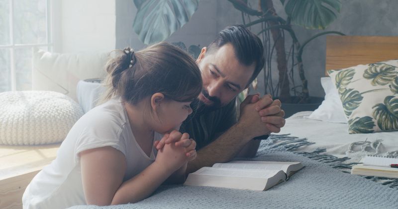 Medium shot of a father and his daughter praying together at home