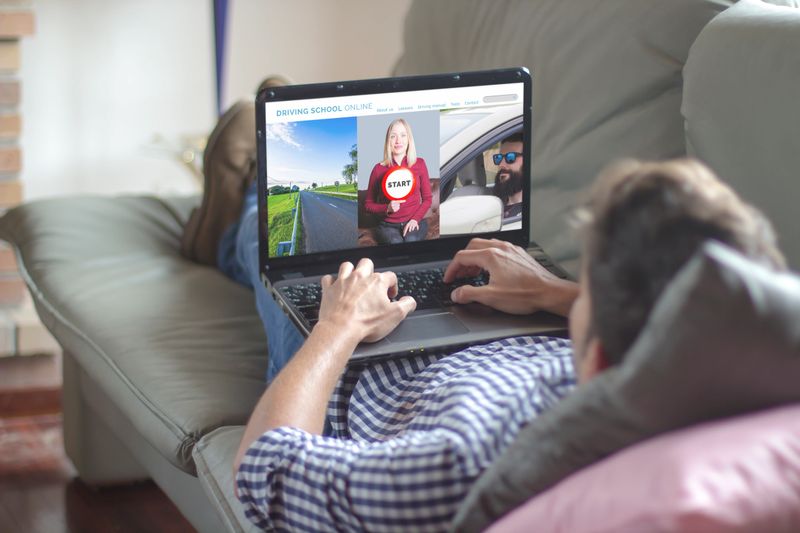 Young man lying comfortably on the sofa at home using laptop with driving school online website on the screen. View from behind.
