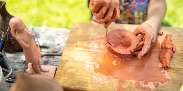 Person sculpting clay pottery outdoors on a wooden board.