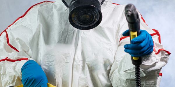 Person in hazmat suit with respirator mask holding a yellow fogging machine.