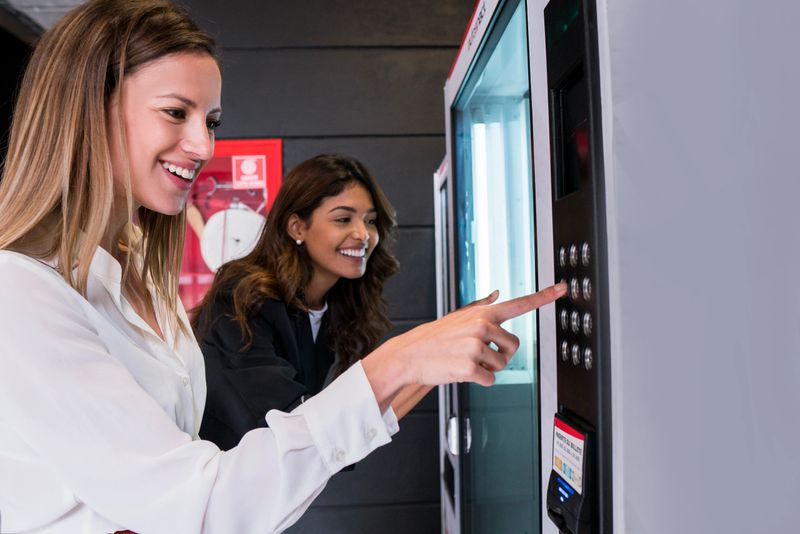 Female coworkers of Latin ethnicity between the ages of 25-35 are on a break from work taking food out of the vending machine in the coworking office