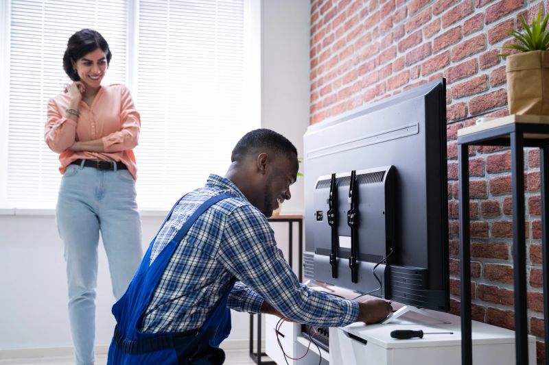 Young Woman Sitting On Couch Looking At Male Technician Repairing TV At Home