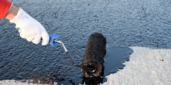 Hand applying black waterproof coating with a roller on concrete surface.