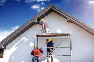 Trabajadores pintando una pared en andamio bajo un cielo azul.