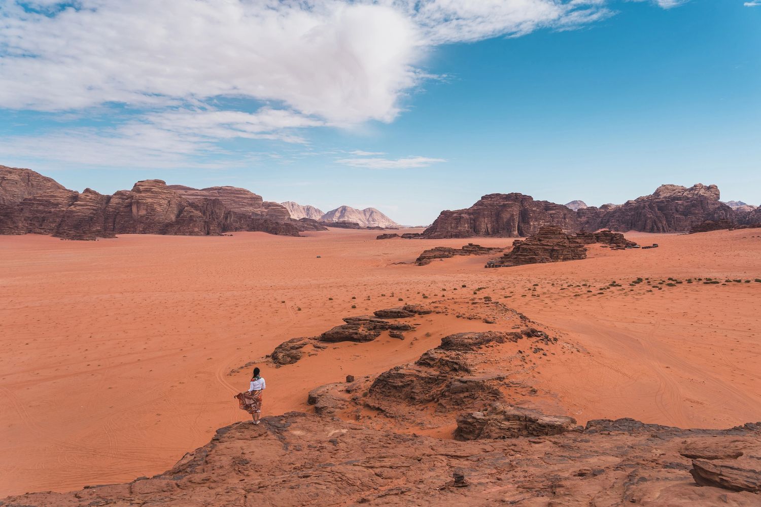 A person stands on rocky terrain overlooking a vast desert with red sand and distant mountains.