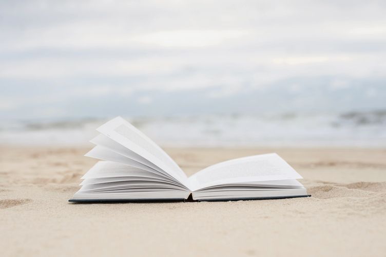 An open book lying on sandy beach with ocean in the background.
