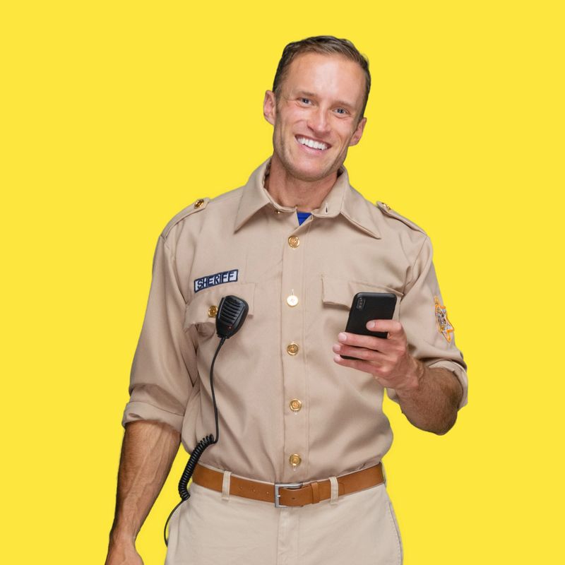 One person of with short hair caucasian male police force standing in front of yellow background wearing badge who is laughing and using smart phone