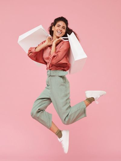 Joyful woman jumping with shopping bags against a pink background.