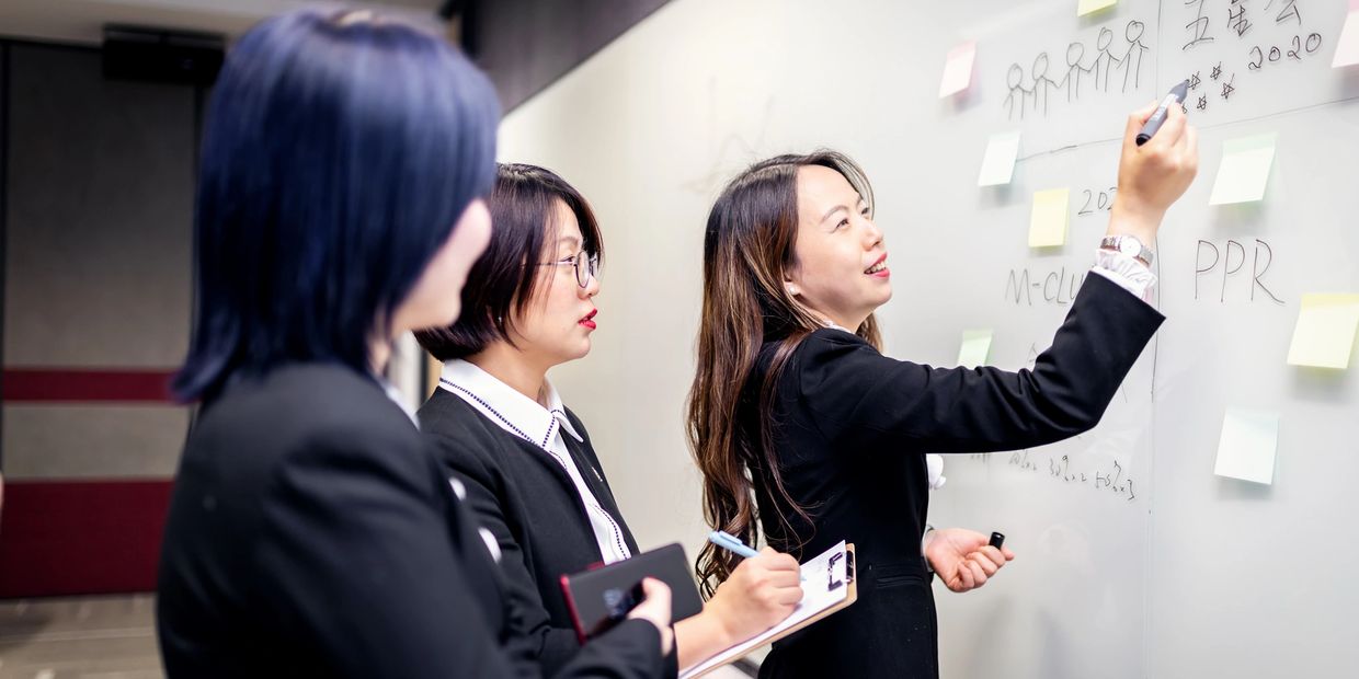 Three professional women collaborating at a whiteboard in an office meeting.
