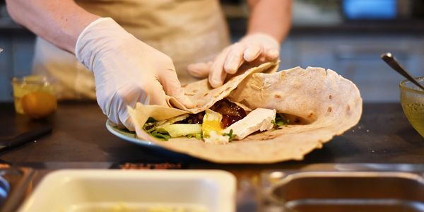 Person with gloves prepares a flatbread wrap with fresh ingredients in a kitchen.