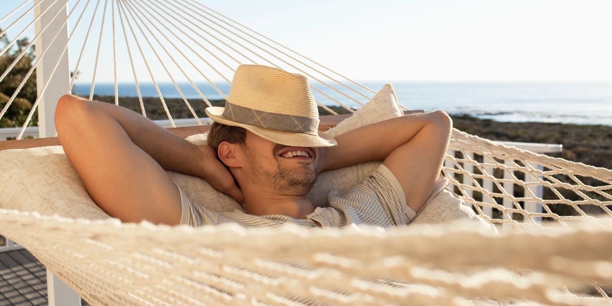 Man relaxing on a hammock by the sea with a hat covering his face.