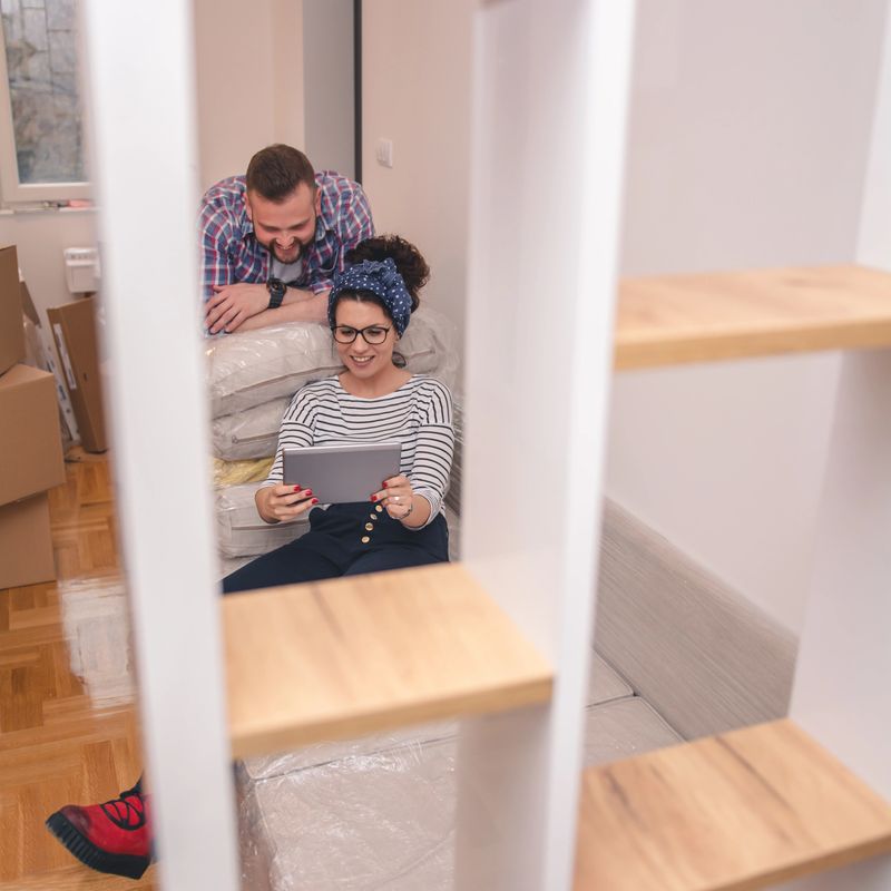 Happy couple using digital tablet sit on sofa with boxes in new home, man and woman renters choose online removal delivery relocation service discuss interior design renovation idea on moving day