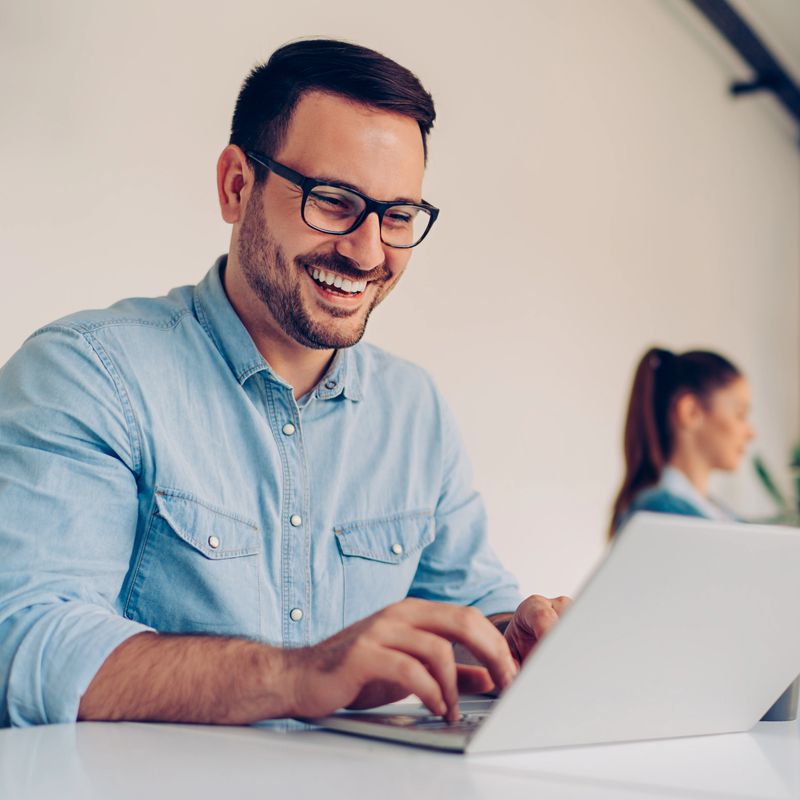 Young businessman sitting by the desk in white office and using laptop with a smile on his face