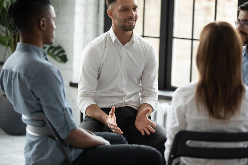 Smiling confident psychologist speaking at group therapy session, young man sharing problems with diverse people sitting in circle, listening to coach at meeting, mentor training staff