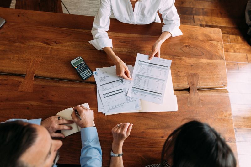 A female financial advisor and accountant meets with a married couple in their home to discuss investments and income. They are sitting at a table discussing savings, investments and taxes with the help of a smart phone and laptop via wireless internet. Image taken in Utah, USA.
