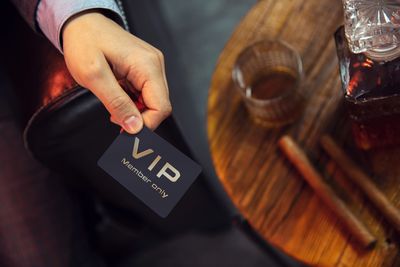 Hand holding a VIP membership card next to a glass of whiskey and cigars.