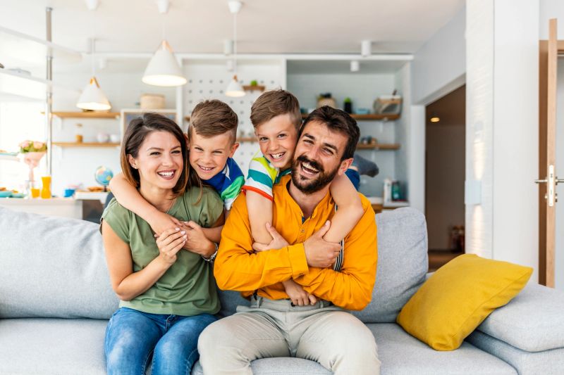 Happy family of four sitting on sofa, looking at camera with big smiles.