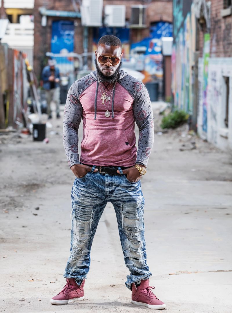 A tough looking African-American man with bling, standing in a city alley, staring at the camera.