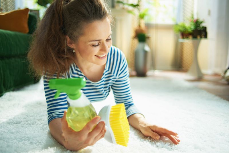 smiling modern middle age housewife in striped t-shirt and white pants with spray bottle of eco friendly cleaning supplies and yellow brush laying on white carpet in the modern house in sunny day.