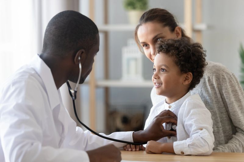 African American male pediatrician with stethoscope listening to lung and heart sound of little boy sitting on mother lap, physician checkup at home or in hospital, children medical insurance care