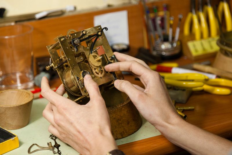 Woman hands fixing with his finger a cogwheel inside a vintage clock mechanism on a workshop unfocused background negative space. Horology business repair concept.