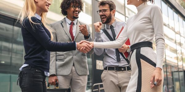 Two women shaking hands while two men cheer outside a modern office building.