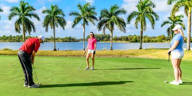 Three people playing golf on a sunny course by a lake with palm trees.