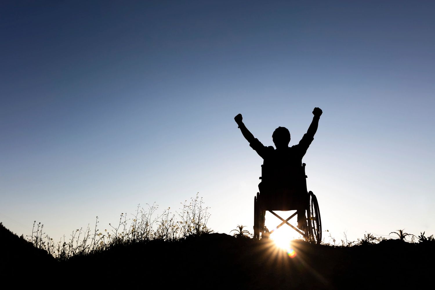 Person in wheelchair raising arms triumphantly at sunrise.