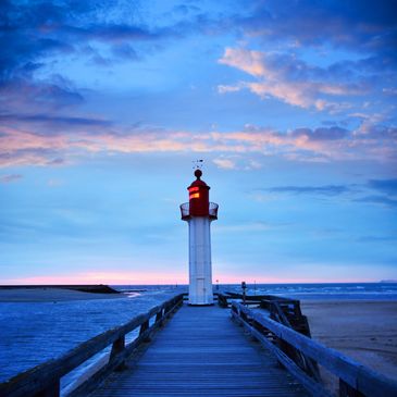 A lighthouse glowing at dusk with a wooden pier leading to it.