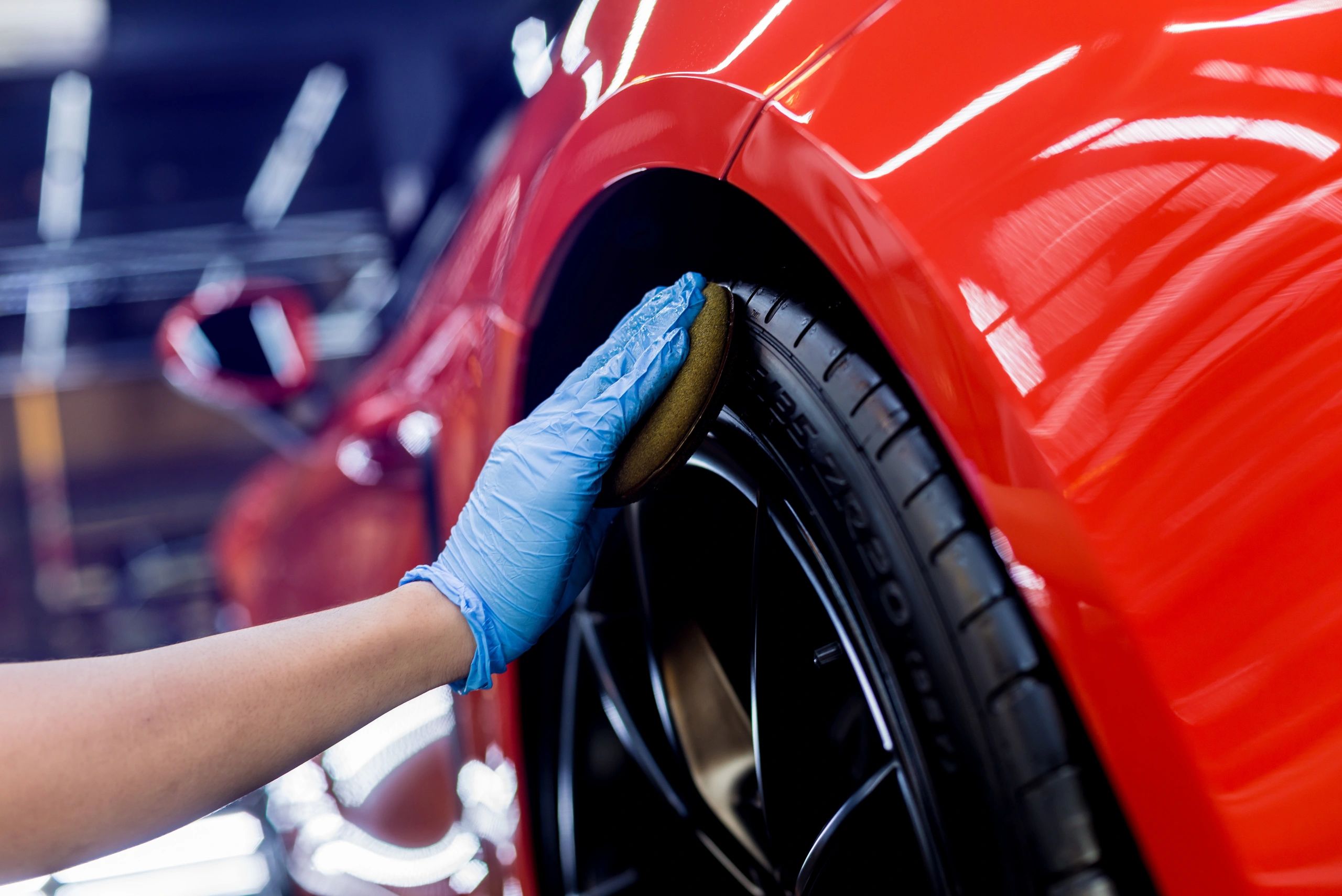 Hand in blue glove polishing a red car's tire with a sponge.