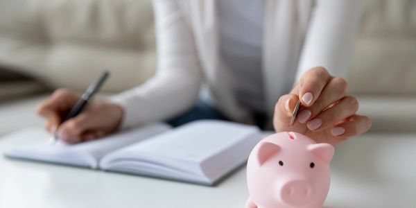 Person saving money by putting a coin in a pink piggy bank while writing in a notebook.