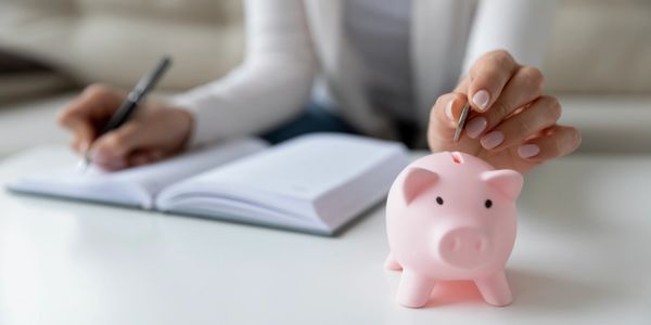 Person saving money by putting a coin in a pink piggy bank while writing in a notebook.