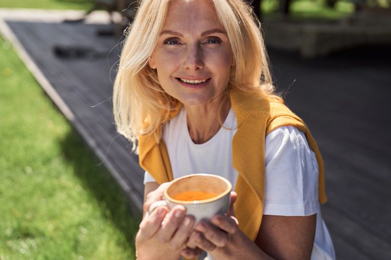 Portrait of smiling beautiful mature female relaxing in countryside yard with hot drink in morning