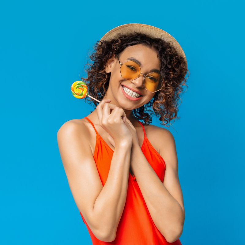 Positive black girl dressed in swimsuit holding sweet lollipop candy and smiling at camera, isolated over blue background with copy space
