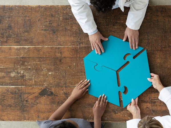 People in white coats assembling large blue puzzle pieces on a wooden table.