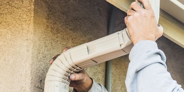 A person installs a beige gutter downspout on a house corner.